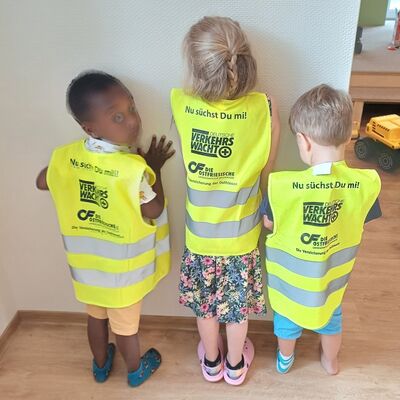 Das Bild zeigt drei Kinder mit dem Rücken zum Betrachter, die jeweils eine Wahrnweste tragen