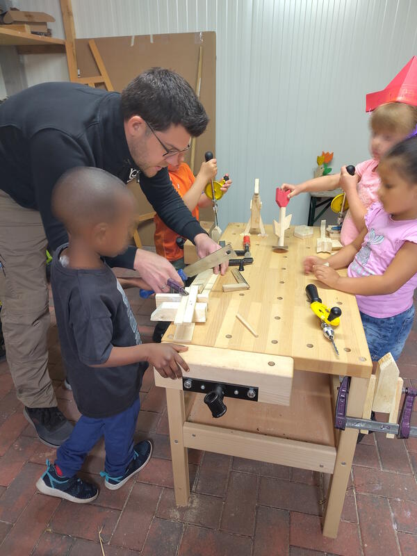 Bild vergr&ouml;&szlig;ern: Das Bild zeigt einen Tischler, der an der Werkbank mit vier Kindern arbeitet