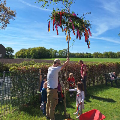 Das Bild zeigt, wie mit den Kindern zusammen der Maibaum aufgestellt wird.