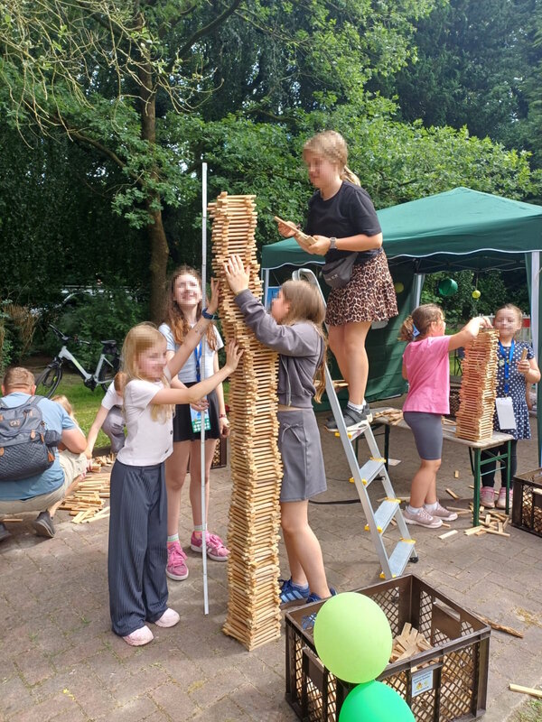 Bild vergr&ouml;&szlig;ern: Das Bild zeigt, wie Kinder mit Leiter und Messstab den aus Holzstäben erbauten Turm vermessen. Er reicht fast an die zwei Meter.