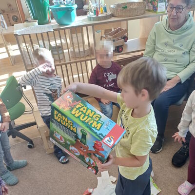 Das Foto zeigt ein Kind im Zentrum des Sitzkreises, wie es ein Tischspiel vom Geschenkpapier befreit und sich freut.