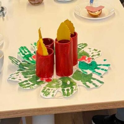 Das Foto zeigt einen aus Pappe, Handabdrücken und Farbe gebastelten Adventskranz auf einem Esstisch in der Upkamer.